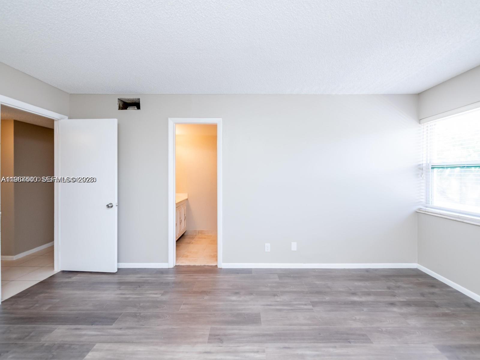An empty room features gray wood-look flooring, light walls, a window, and two doorways — 2304 NE 7th St # 27, Hallandale Beach FL 33009, HALLANDALE BEACH, FL