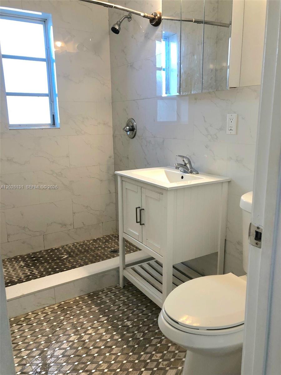 A bathroom features a white vanity, a toilet, and a shower with marble-look wall tiles — 7135 Rue Granville # 4, Miami Beach FL 33141, MIAMI BEACH, FL