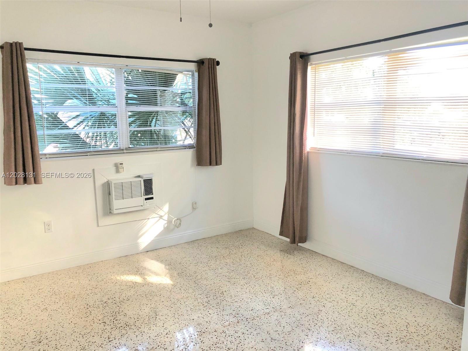 An empty room features white walls, terrazzo flooring, two windows with curtains, and an air conditioner — 7135 Rue Granville # 4, Miami Beach FL 33141, MIAMI BEACH, FL