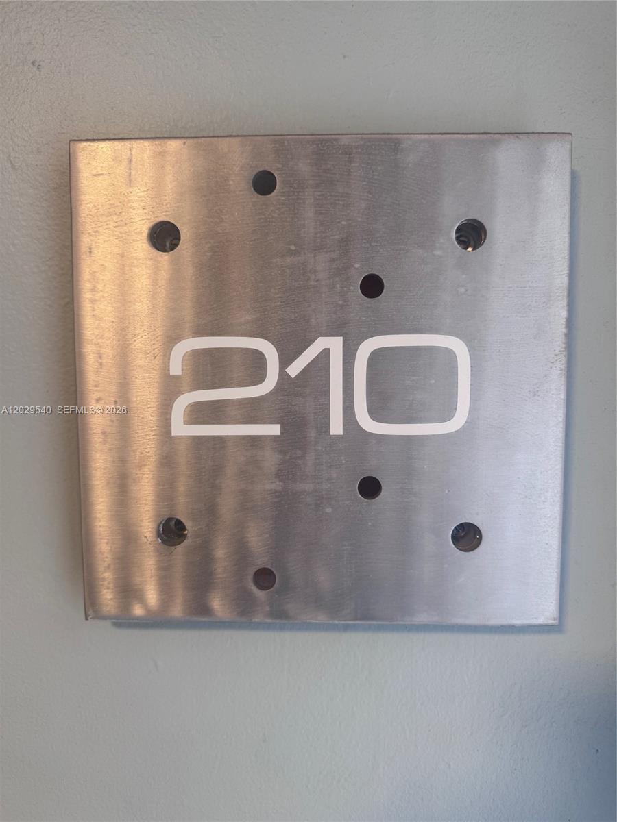 A brushed metal square sign displays the white number 210 with several circular cutouts — 865 Collins Ave # 210, Miami Beach FL 33139, MIAMI BEACH, FL