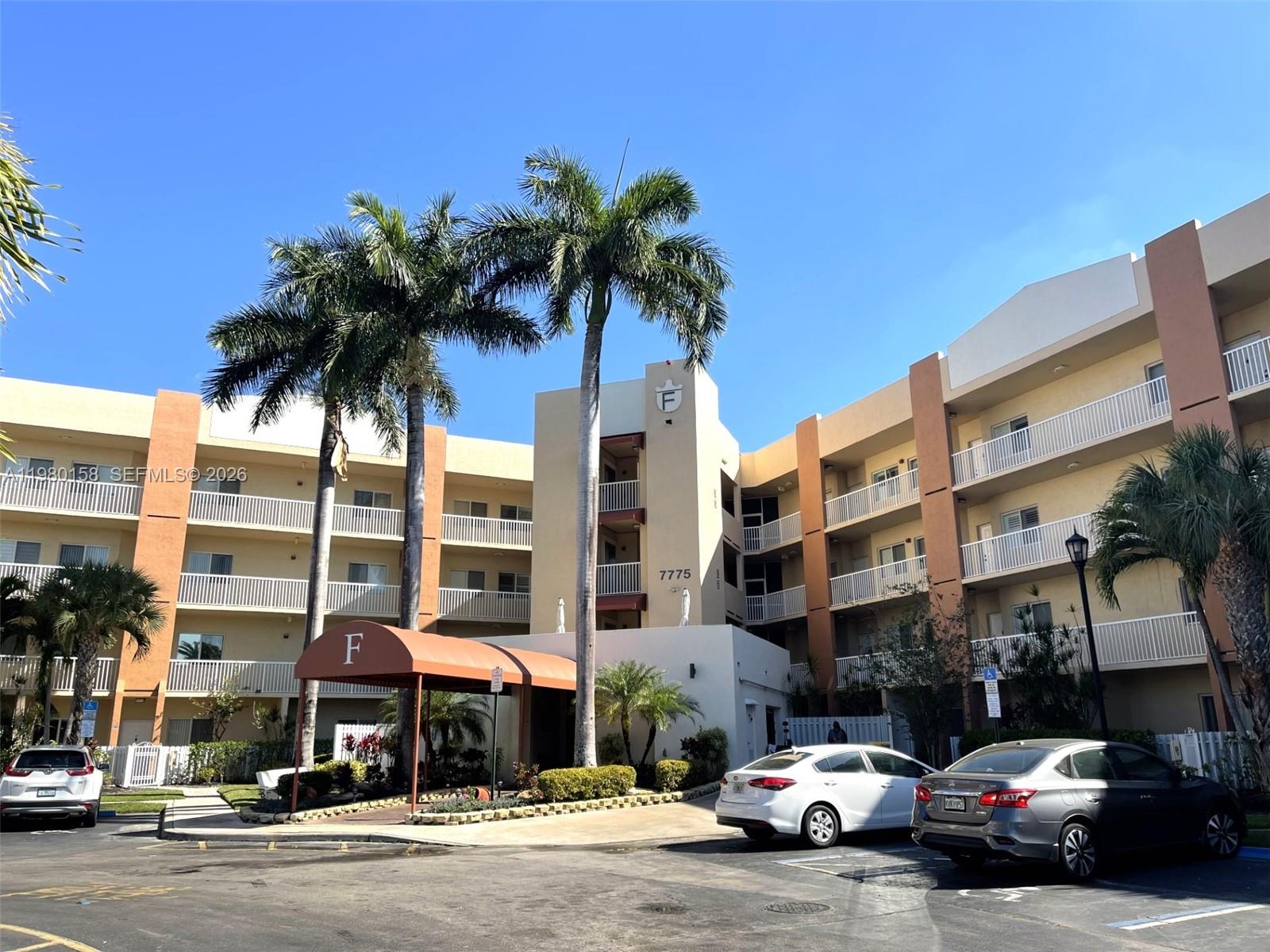 The exterior of a multi-story apartment building features palm trees and a covered entrance — 7775 Yardley Dr # 409, Tamarac FL 33321, TAMARAC, FL
