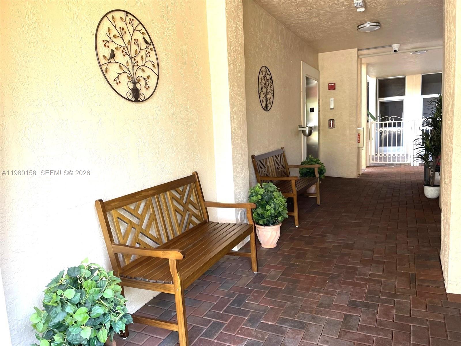 An outdoor covered walkway features brick flooring, two wooden benches, and decorative wall art — 7775 Yardley Dr # 409, Tamarac FL 33321, TAMARAC, FL