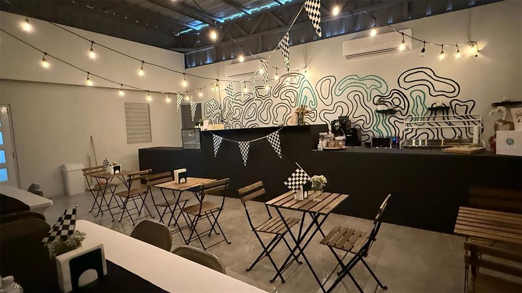 A cafe interior features small wooden tables, string lights, and a black service counter — 156 SECTOR, KM 13.5 PR-156, BARRANQUITAS, PR