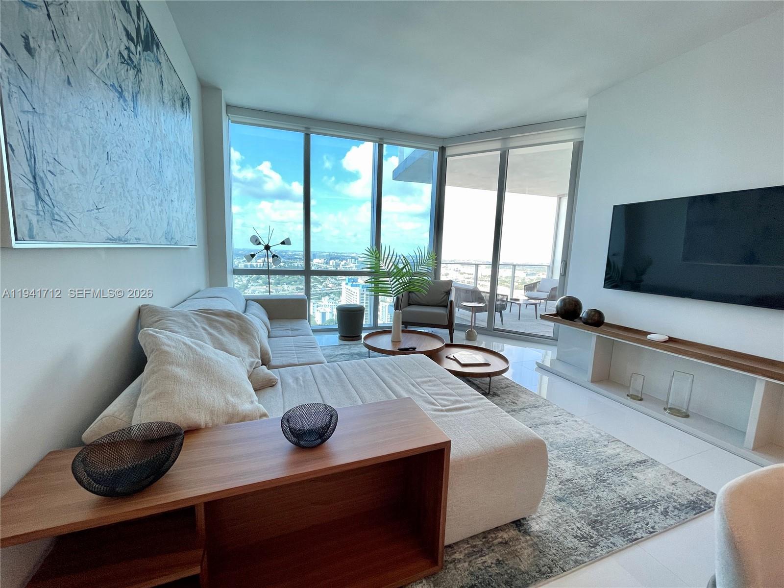 A modern living room features a sectional sofa, wooden coffee tables, and floor-to-ceiling windows — 851 NE 1st Ave # 3606, Miami FL 33132, MIAMI, FL