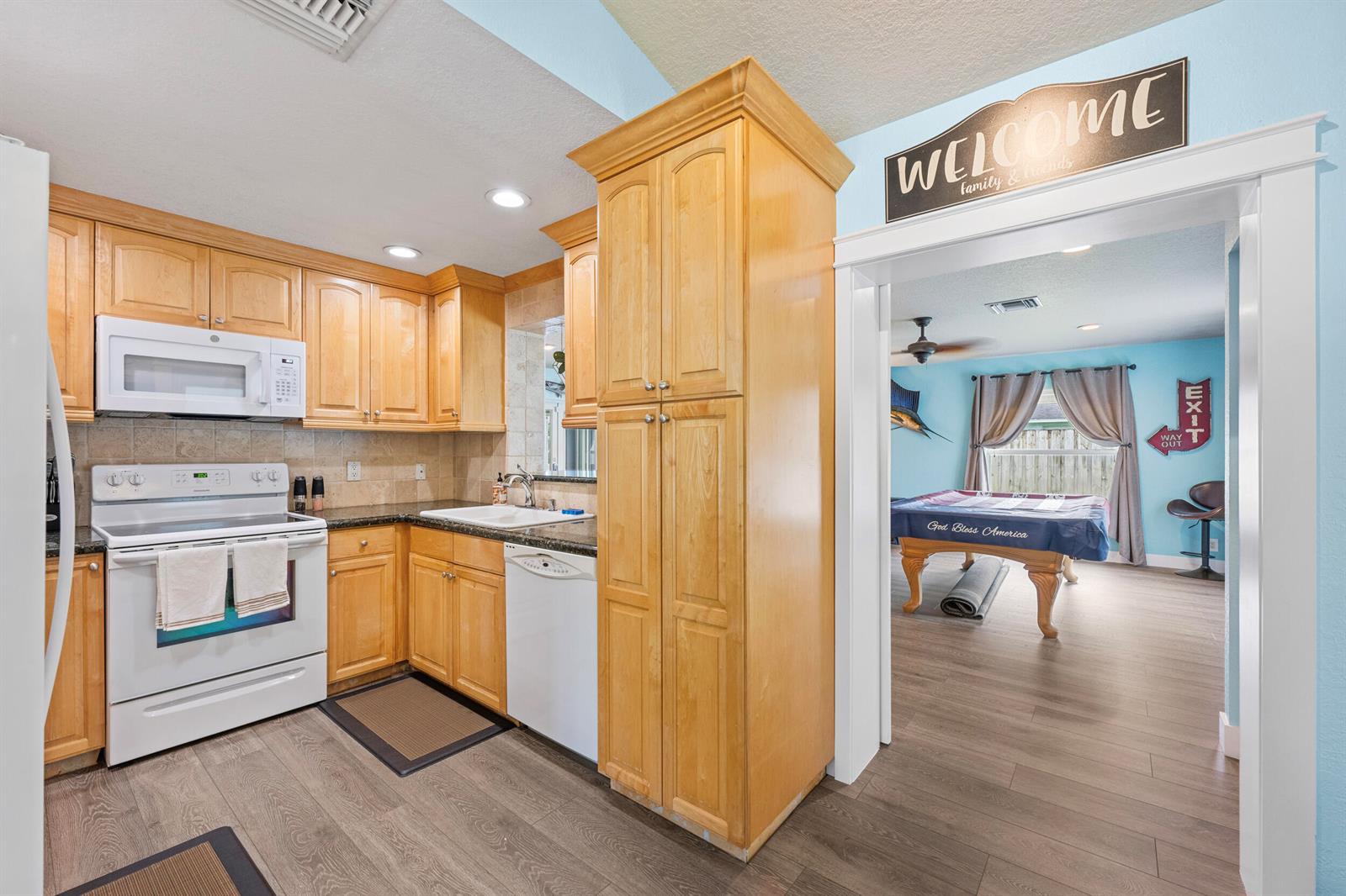 A kitchen with light wood cabinets and white appliances opens into a game room — 128 Banyan Circle, Jupiter FL 33458, JUPITER, FL