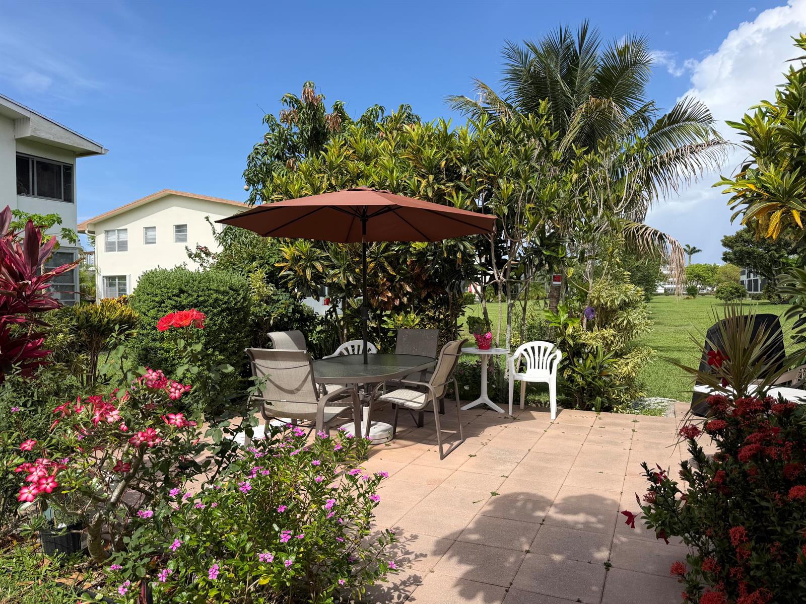 A patio features a dining set with a brown umbrella, surrounded by lush garden landscaping — 162 Windsor H, West Palm Beach FL 33417, WEST PALM BEACH, FL