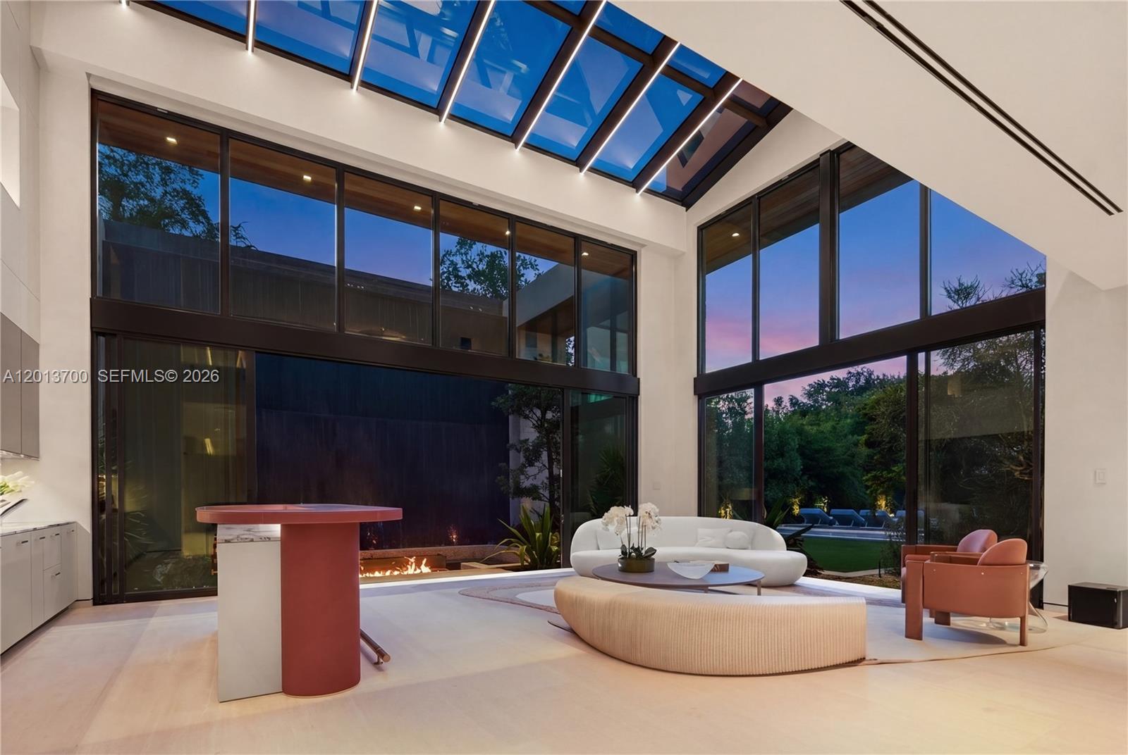 A modern living room features a glass skylight, floor-to-ceiling windows, and a central fireplace — 6701 SW 101st St, Pinecrest FL 33156, PINECREST, FL