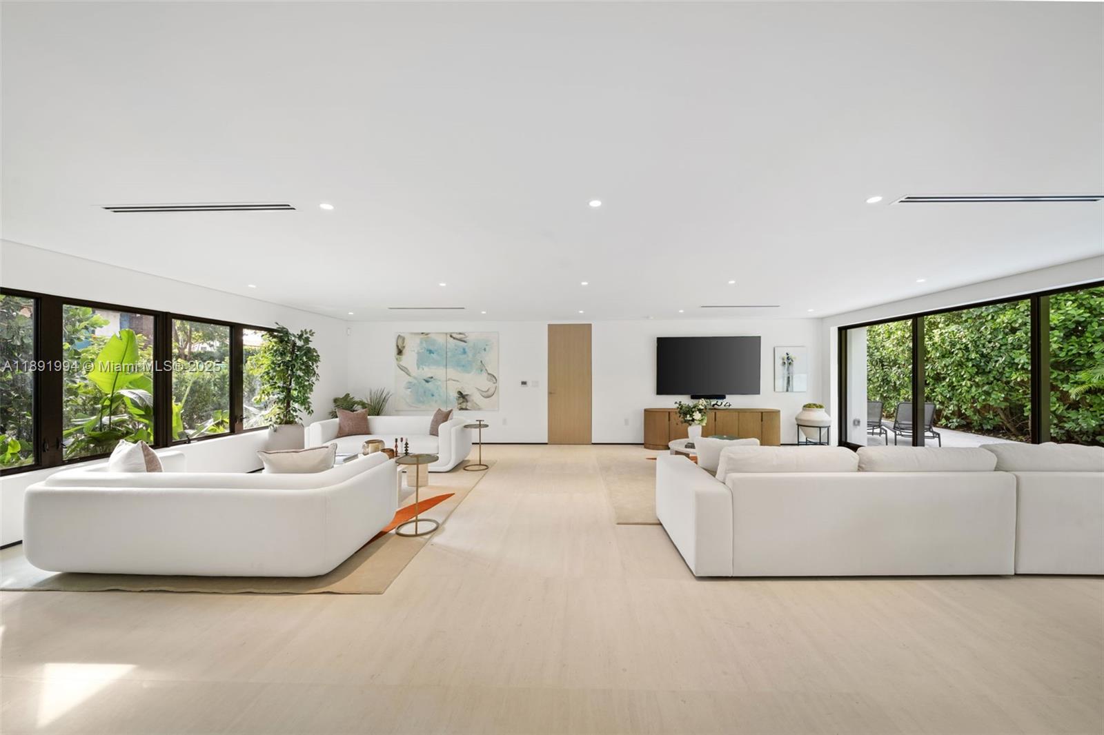 A bright living room features white sofas, light hardwood floors, and large windows overlooking greenery — 222 E Rivo Alto Dr, Miami Beach FL 33139, MIAMI BEACH, FL