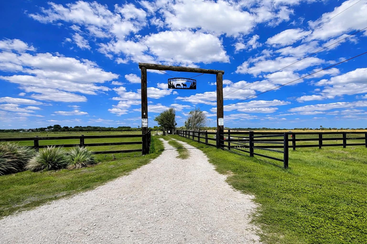 A gravel driveway leads through a wooden gate entrance into a fenced pasture under blue skies — 8121 NW 58th Lane, Okeechobee FL 34972, OKEECHOBEE, FL