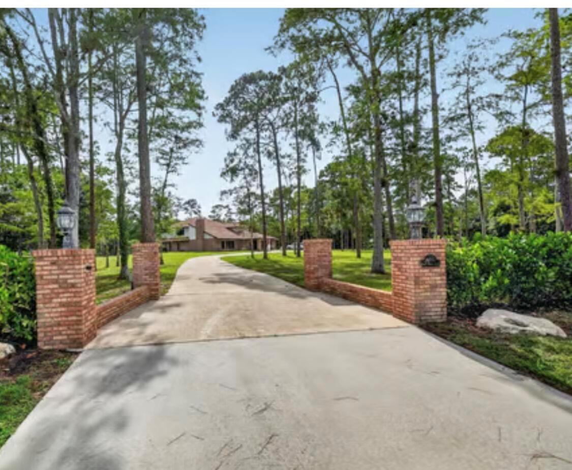 A concrete driveway leads through brick entrance pillars toward a house surrounded by tall trees — 14780 Draft Horse Lane, Wellington FL 33414, WELLINGTON, FL