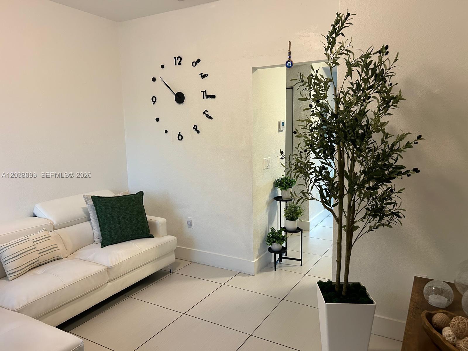 A living room features a white sofa, a large wall clock, and a potted olive tree — 106 SE 9th Ct # 201, Hallandale Beach FL 33009, HALLANDALE BEACH, FL
