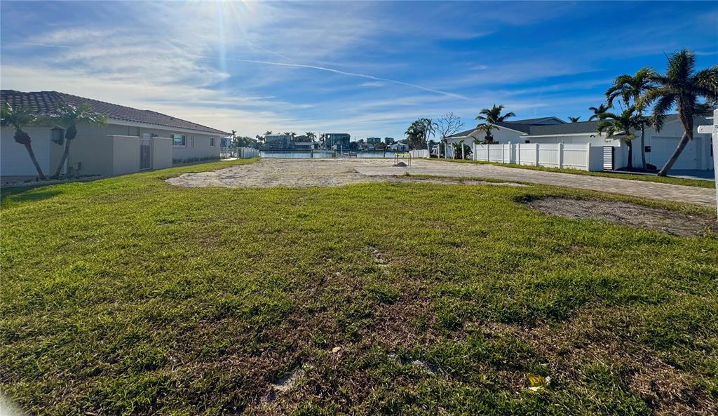 A vacant waterfront lot features a grassy foreground, sandy clearing, and surrounding residential properties — 7874 9TH AVENUE S, ST PETERSBURG, FL