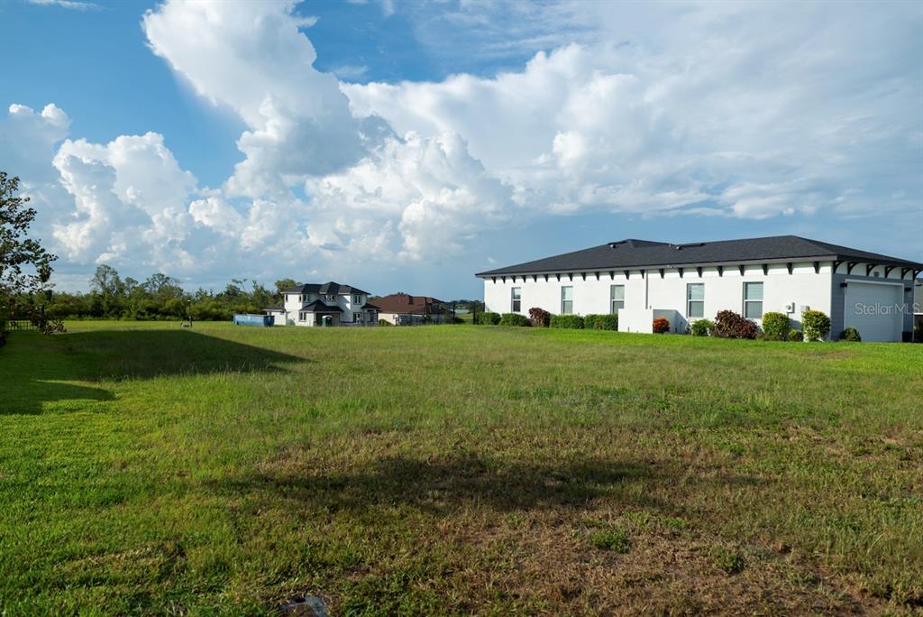 A large, grassy vacant lot sits in front of a modern white house under clouds — 338 SPINNAKER COURT, AUBURNDALE, FL