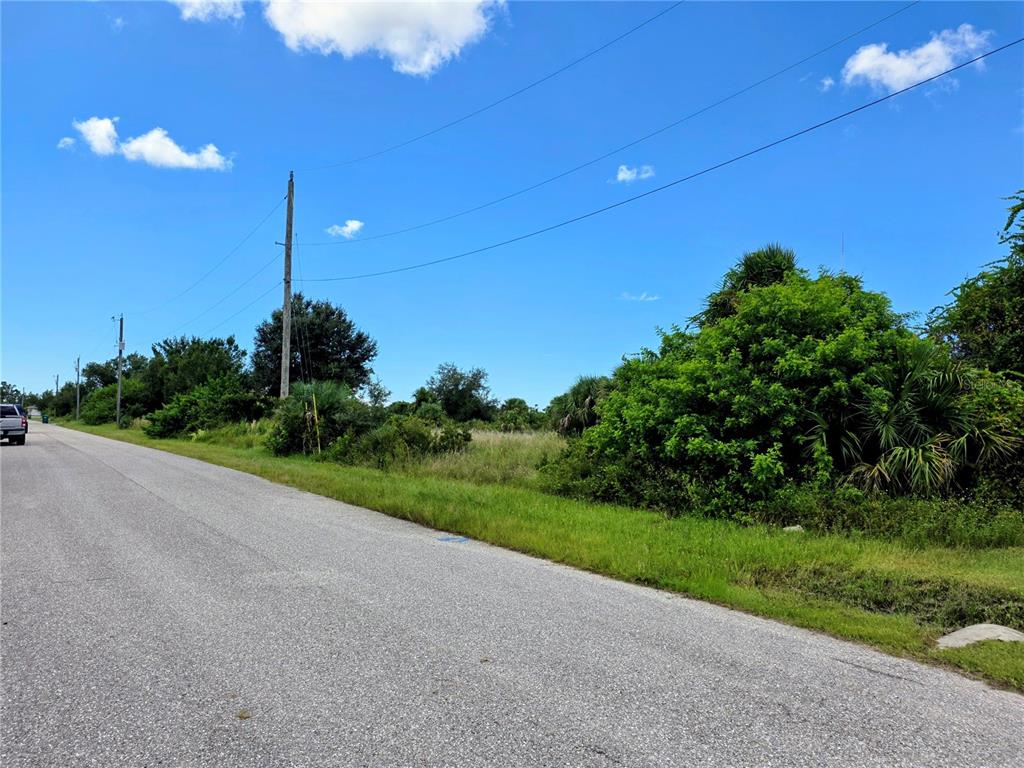 A paved road runs alongside a vacant, overgrown lot under a bright blue sky — 9177 ARBERG STREET, PORT CHARLOTTE, FL