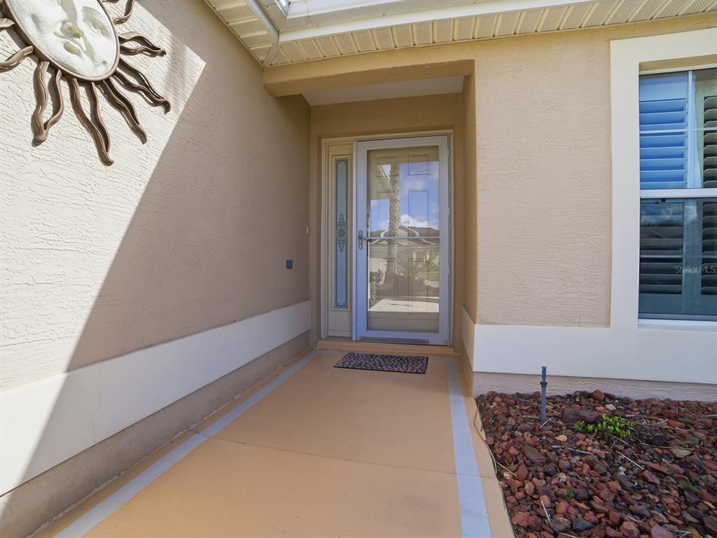 A tan exterior entryway features a storm door, a sun wall decoration, and a window — 1104 ALCOVE LOOP, THE VILLAGES, FL