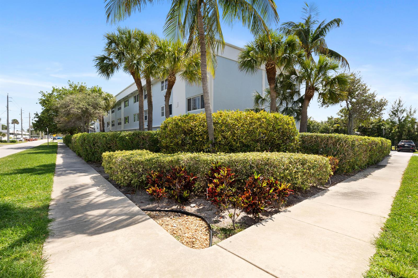 A multi-story apartment building sits behind a manicured hedge and palm trees near sidewalks — 505 NE 20th Avenue # 112, Deerfield Beach FL 33441, DEERFIELD BEACH, FL