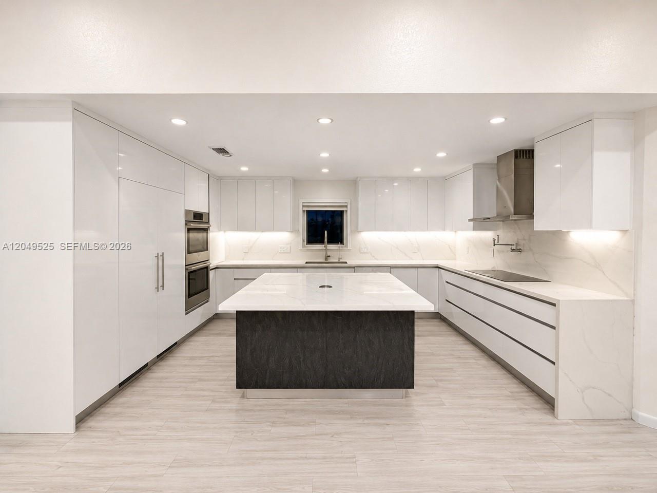 A modern kitchen features white cabinetry, a dark central island, and light wood flooring — 2616 Oakbrook Ct, Weston FL 33332, WESTON, FL