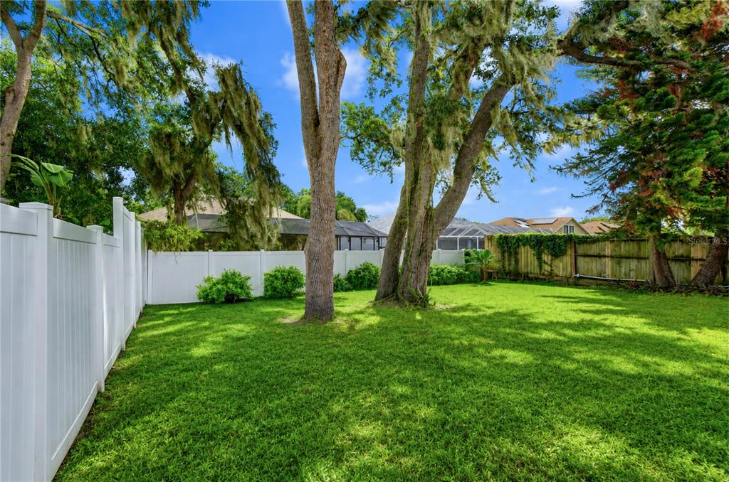A grassy backyard features two large trees and is enclosed by white vinyl and wooden fencing — 2512 WRENCREST CIRCLE, VALRICO, FL