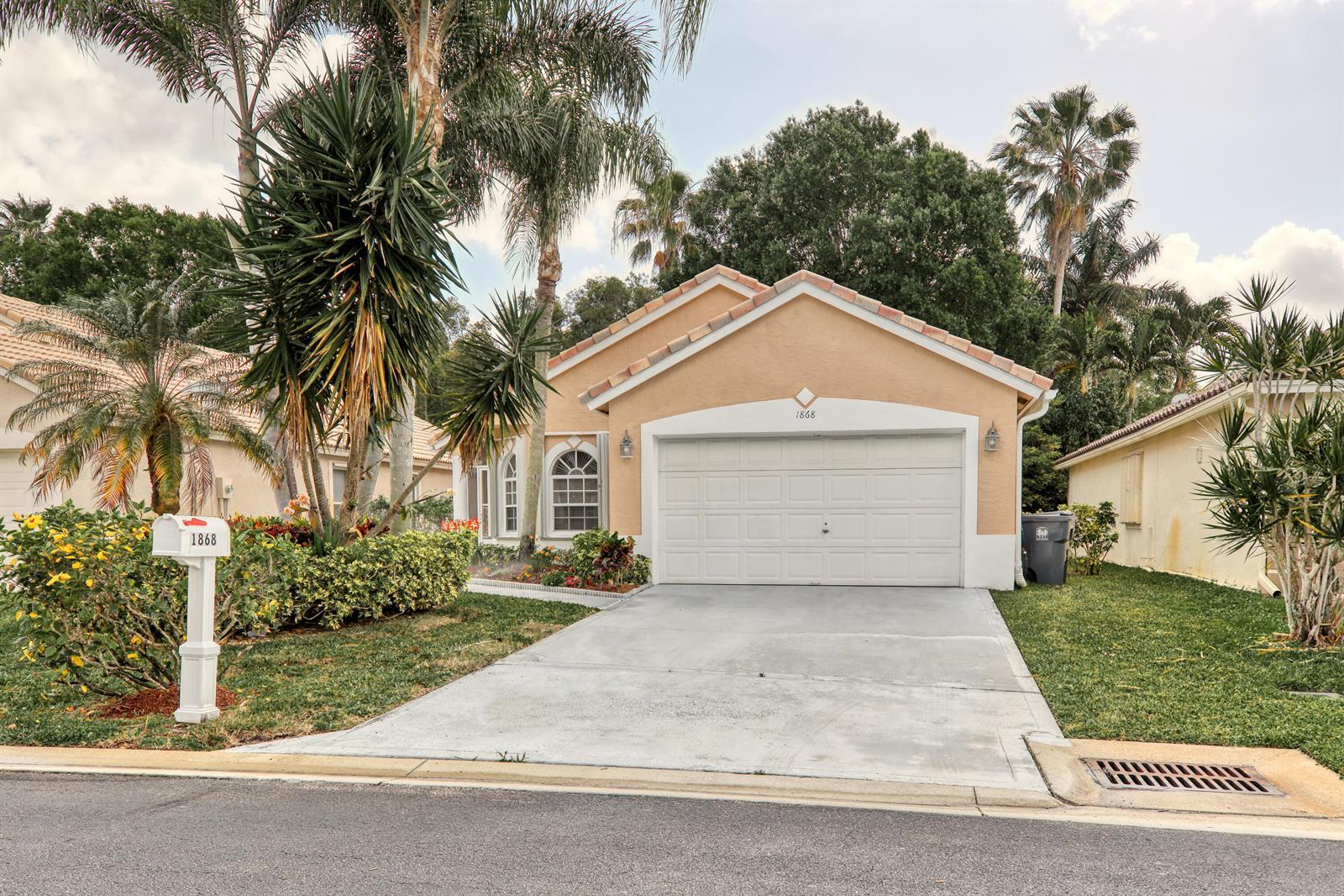 A single-story tan house with a white garage door and a palm-filled front yard — 1868 Barnstable Road, Wellington FL 33414, WELLINGTON, FL