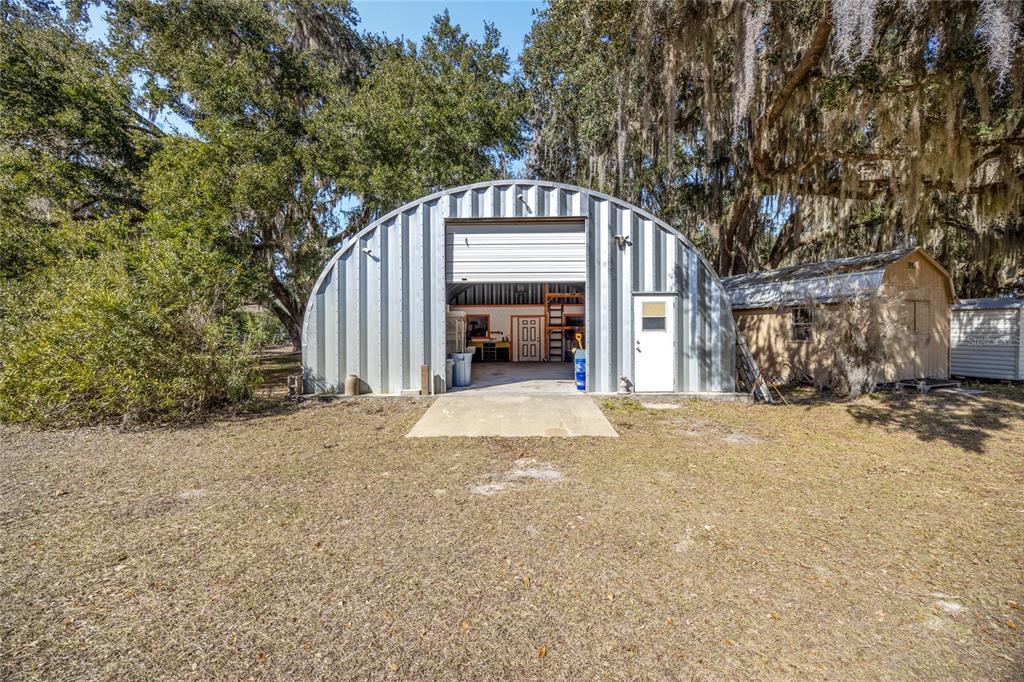 A metal Quonset hut workshop stands in a grassy yard under large oak trees — 16602 NE 124TH AVENUE, WALDO, FL
