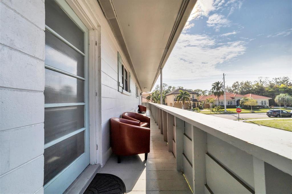 A long, covered balcony features light-colored wood flooring, a blue door, and two brown chairs — 1901 HYDE PARK STREET UNIT C, SARASOTA, FL