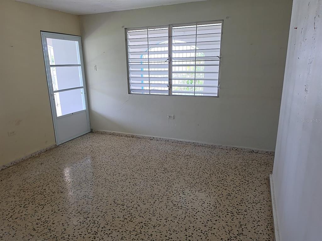 An empty room features light-colored walls, a window with metal louvers, and speckled tile flooring — 8 CALLE CASCADA, SAN LORENZO, PR