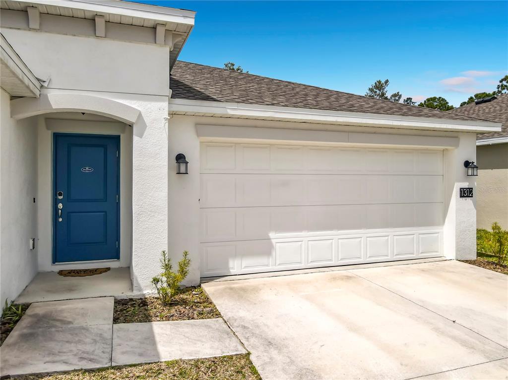 A white house exterior features a blue front door and a two-car garage — 1312 PEACH CREEK DRIVE, OSTEEN, FL