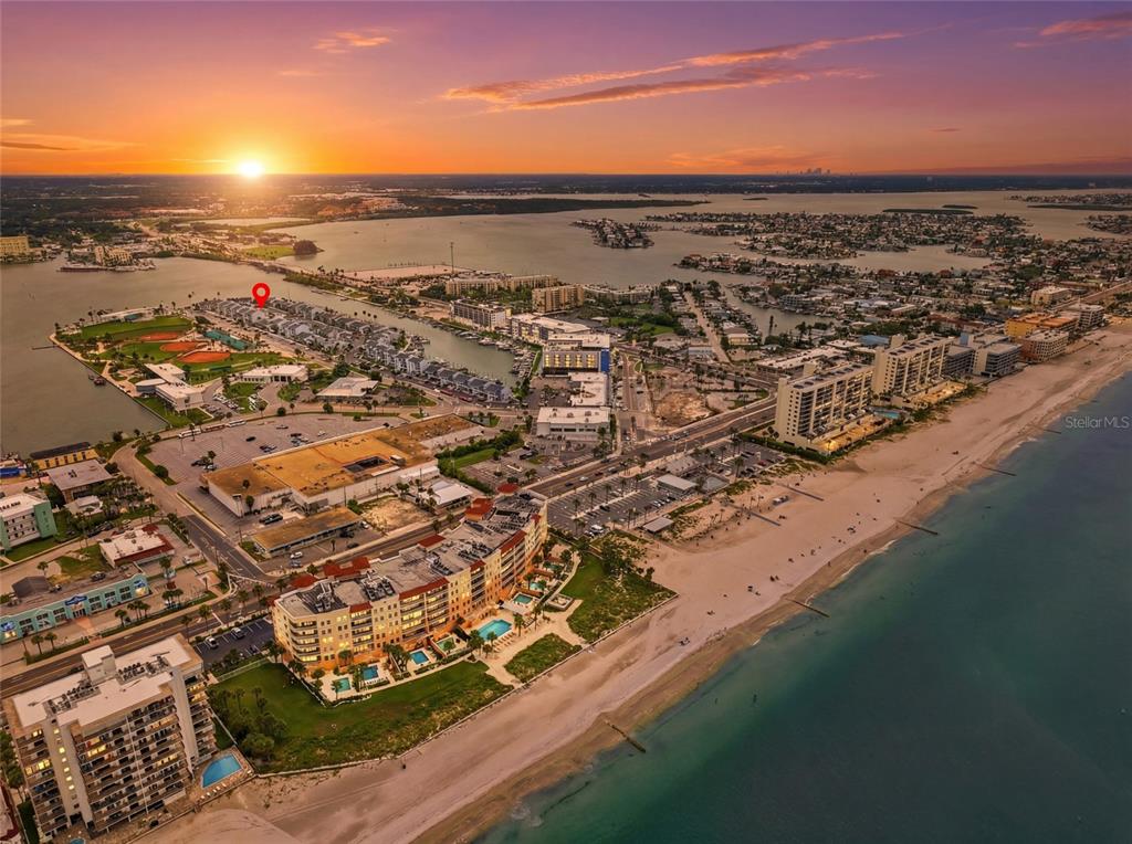 An aerial view shows a coastal town with a beach, buildings, and a sunset horizon — 320 MEDALLION BOULEVARD UNIT H, MADEIRA BEACH, FL