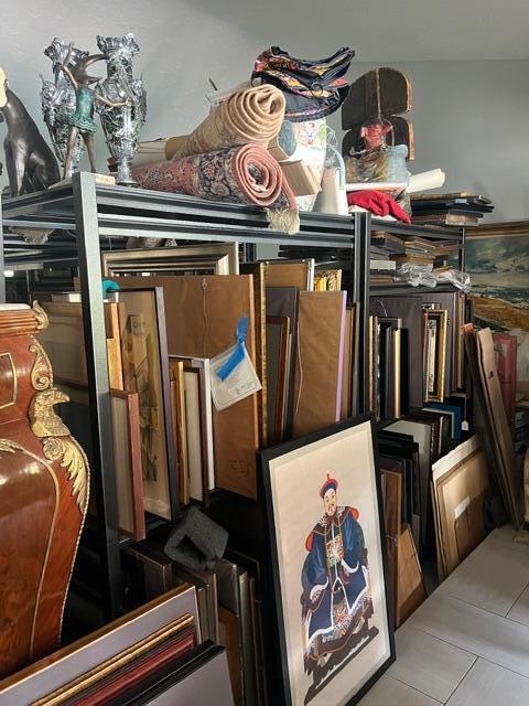 A metal shelving unit is packed with various framed artworks, rolled rugs, and decorative objects — Wellington Trace, Wellington FL, WELLINGTON, FL