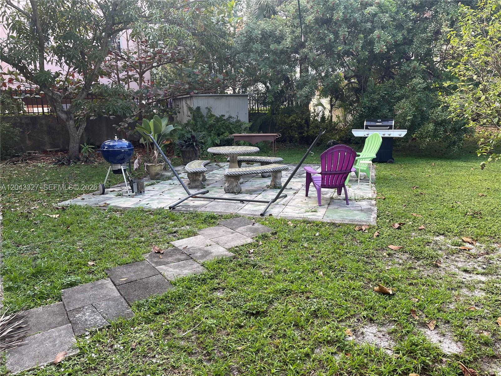 A backyard patio features a stone table, benches, a hammock stand, and two colorful chairs — CORAL GABLES, FL