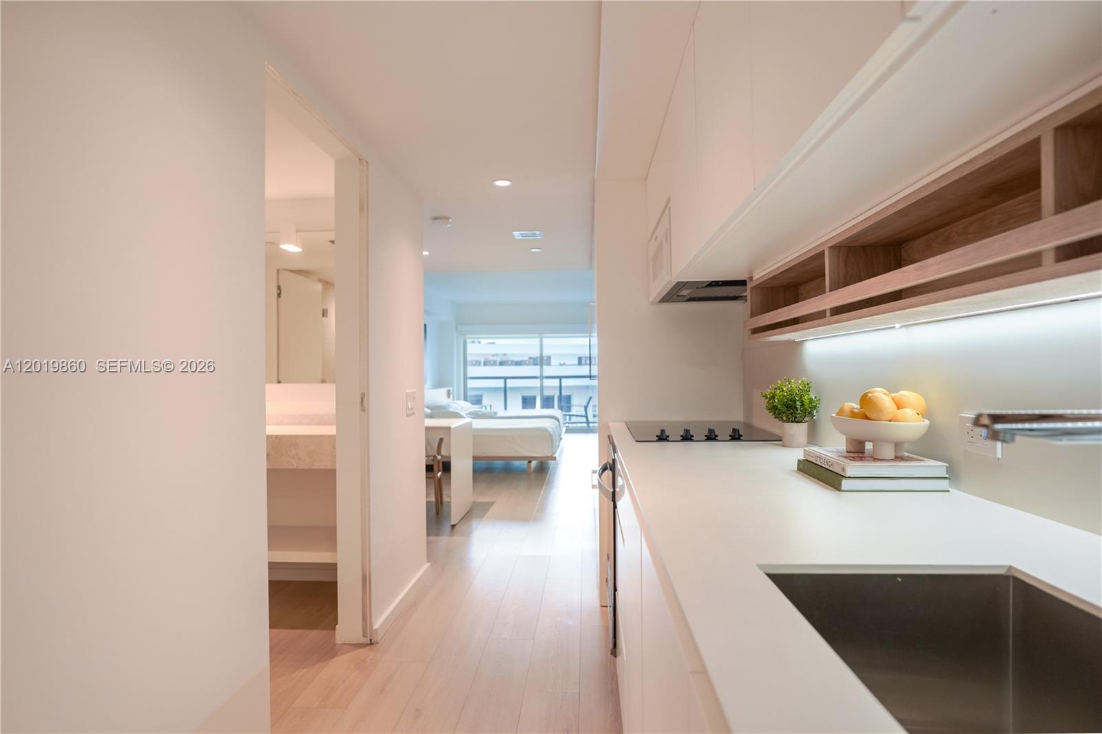 A modern kitchen with white countertops and cabinetry leads into a bedroom with a balcony — 10250 Collins Avenue # 407, Bal Harbour FL 33154, BAL HARBOUR, FL