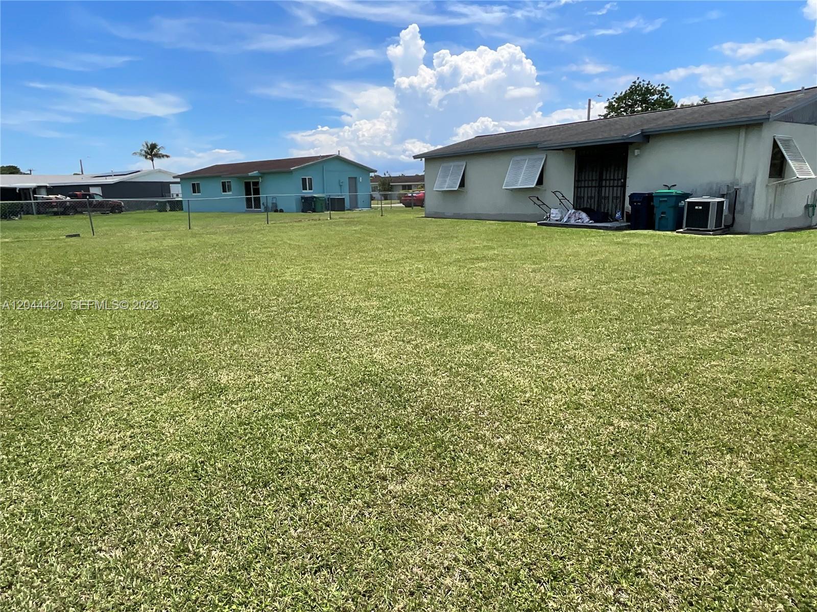 A large grassy backyard sits between two single-story houses under a blue sky — 14253 SW 107th Pl # X, Miami FL 33176, MIAMI, FL
