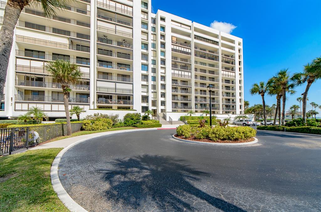 A multi-story white apartment building features balconies and a circular driveway with landscaping — 1460 GULF BOULEVARD UNIT 1105, CLEARWATER BEACH, FL