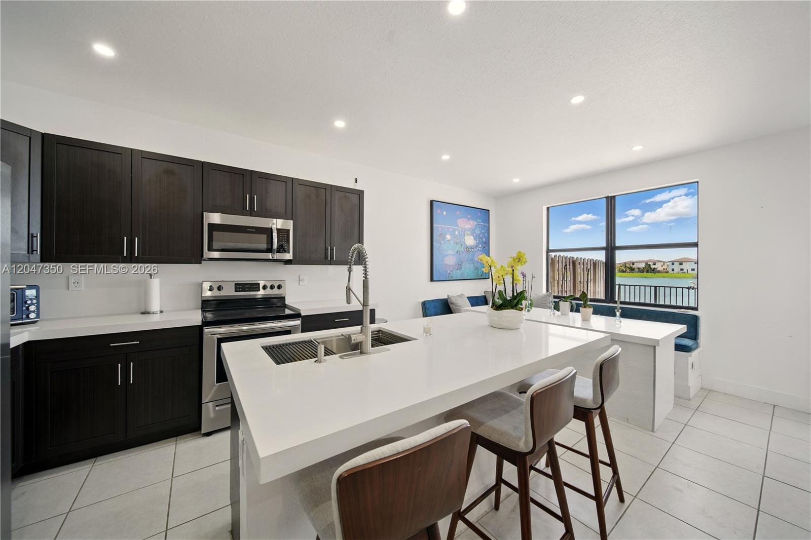 A modern kitchen features dark cabinetry, a white island, and a dining nook overlooking water — 10269 SW 231st Ln # 0, Miami FL 33190, MIAMI, FL