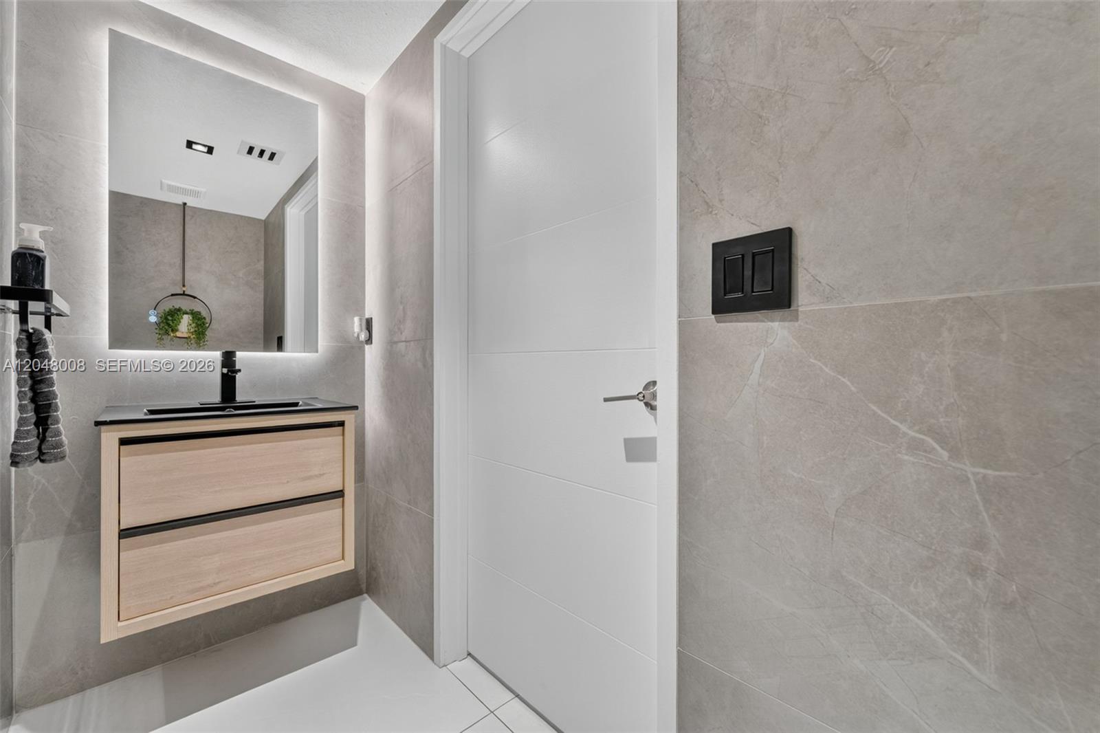 A modern bathroom features a floating wood vanity, a backlit mirror, and gray tiled walls — 11024 W 35th Ln, Hialeah FL 33018, HIALEAH, FL
