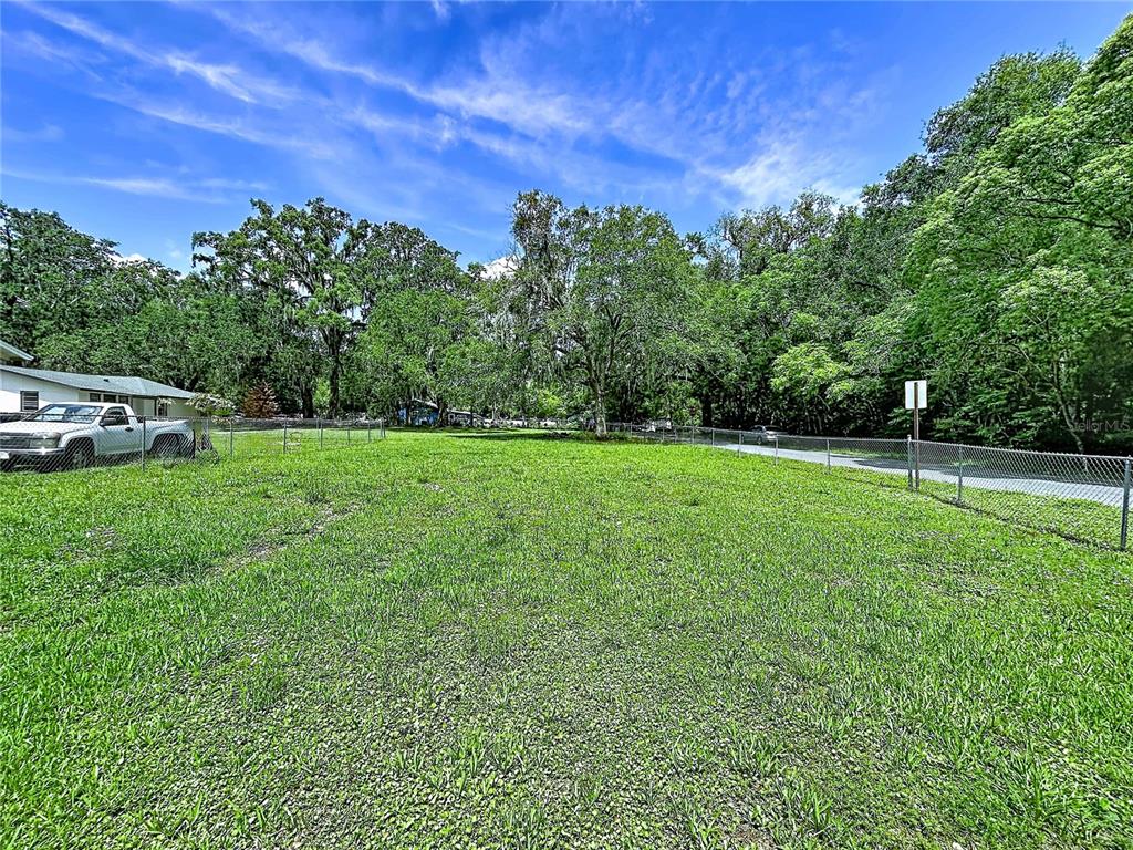 A grassy, fenced-in vacant lot is surrounded by mature trees under a blue sky — 0 EAST AVE, BROOKSVILLE, FL