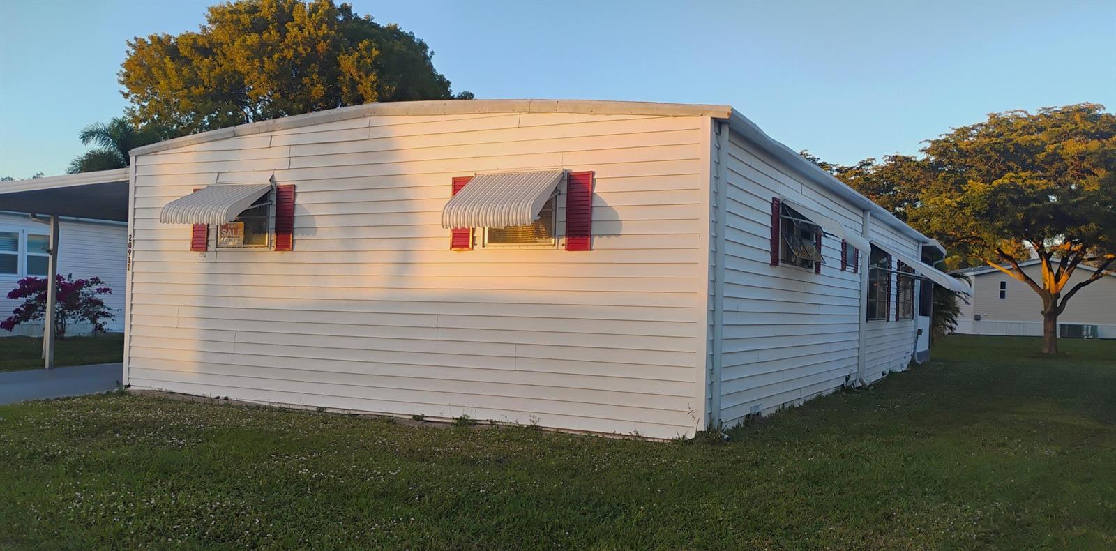 A white mobile home with red shutters and metal window awnings sits on a grassy lot — 20911 Sunrise Drive, Boca Raton FL 33433, BOCA RATON, FL