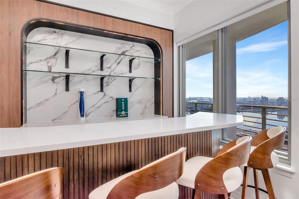 A modern home bar features a marble backsplash, glass shelving, and wood-paneled seating by windows — 1209 E CUMBERLAND AVENUE UNIT 1701, TAMPA, FL