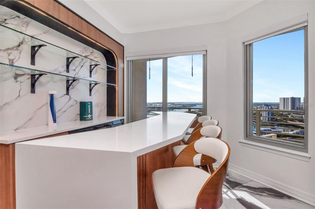 A modern home bar features a white countertop, wood cabinetry, glass shelving, and waterfront views — 1209 E CUMBERLAND AVENUE UNIT 1701, TAMPA, FL