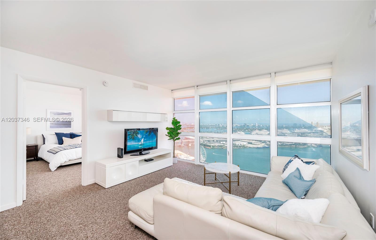A living room with a white sectional sofa, television, and floor-to-ceiling windows overlooking the water — 50 Biscayne Blvd # 4104, Miami FL 33132, MIAMI, FL