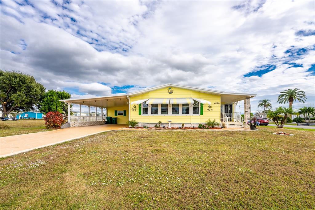 A yellow manufactured home with a carport and a front lawn under a cloudy sky — 6290 GREENFINCH ROAD, ENGLEWOOD, FL