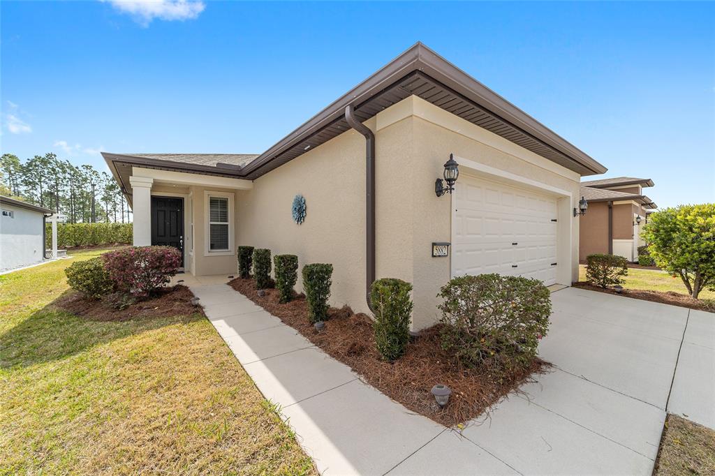 A tan stucco house features a single-car garage, a front walkway, and landscaped bushes — 5882 SW 93RD CIRCLE, OCALA, FL