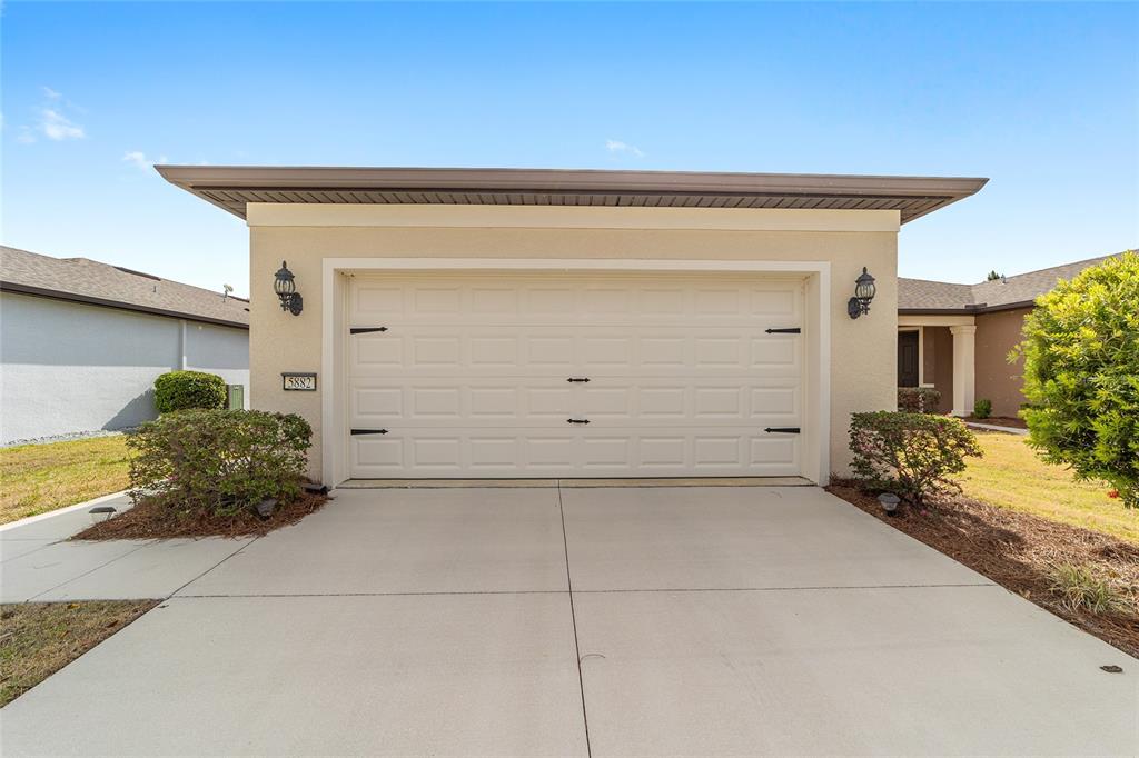 A beige house exterior features a two-car garage with a white door and concrete driveway — 5882 SW 93RD CIRCLE, OCALA, FL
