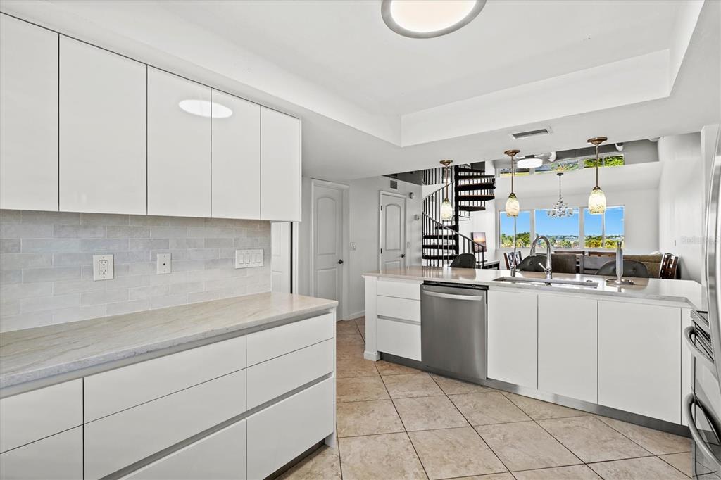 A modern white kitchen features a stainless steel dishwasher, tile flooring, and a spiral staircase — 9604 CORTEZ ROAD W UNIT 134, BRADENTON, FL