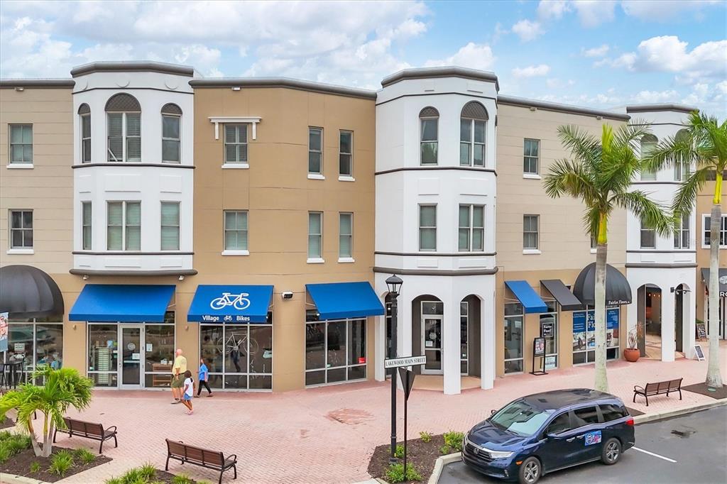 A multi-story commercial building with retail storefronts, blue awnings, and a parking area in front — 8111 LAKEWOOD MAIN STREET UNIT 306, LAKEWOOD RANCH, FL