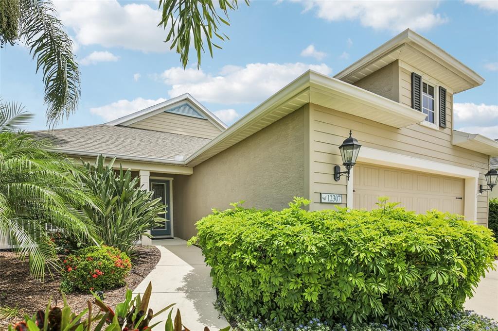 A tan house exterior features a front walkway, a garage, and lush landscaping — 12307 WHISPER LAKE DRIVE, BRADENTON, FL