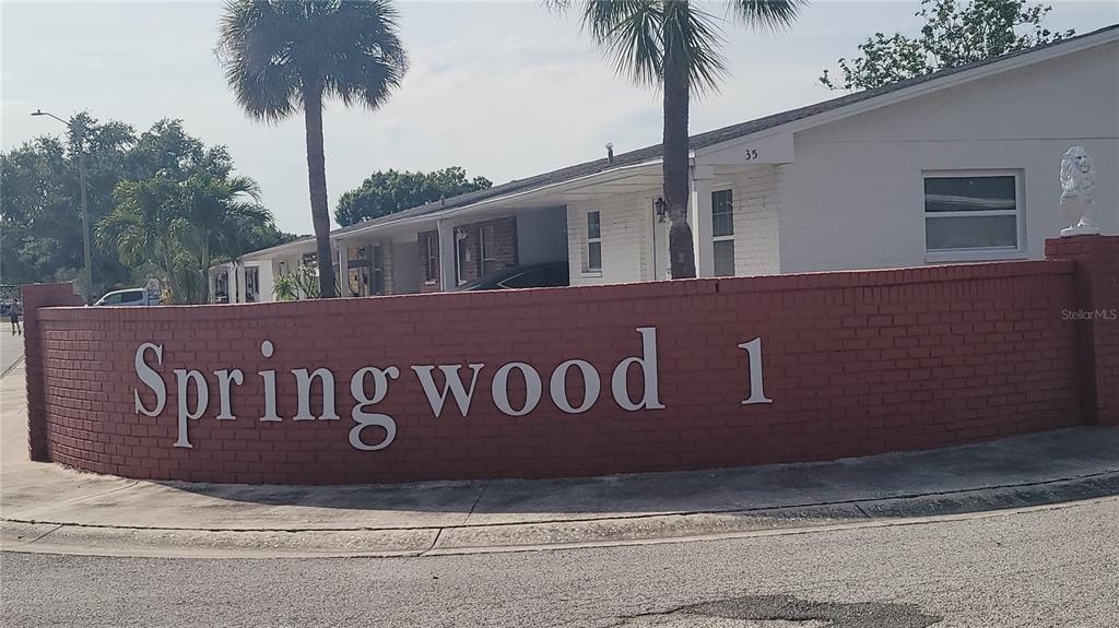 A curved red brick wall displays the name Springwood 1 in front of a white building — 9850 LILY STREET N UNIT 117, PINELLAS PARK, FL