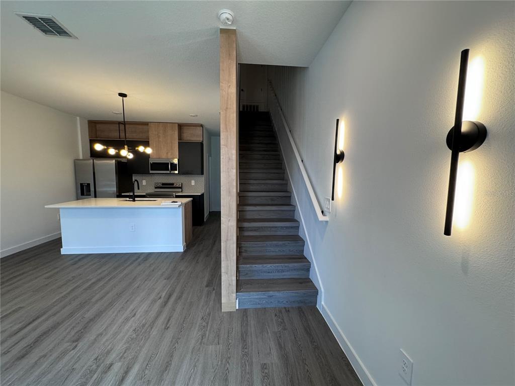 A modern kitchen with a white island sits beside a staircase with wood-look flooring — 3239 SUSTAINABLE WAY, KISSIMMEE, FL