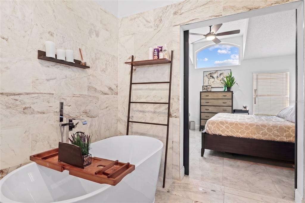 A bathroom with a freestanding tub and marble tile walls opens into a bedroom — 6847 BOUGANVILLEA CRESCENT DRIVE, ORLANDO, FL