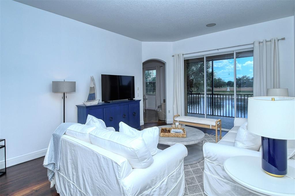 A living room features white sofas, a blue media console, and sliding doors overlooking water — 8363 MIRAMAR WAY UNIT 203, LAKEWOOD RANCH, FL